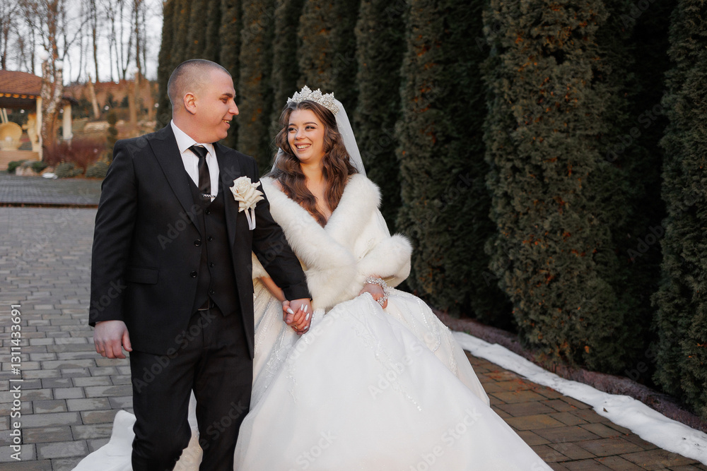 A bride and groom are walking down a path in a snowy setting. The bride is wearing a white dress and a fur stole, while the groom is wearing a suit and tie. They are holding hands and smiling A bride and groom are walking down a path in a snowy setting. The bride is wearing a white dress and a fur stole, while the groom is wearing a suit and tie. They are holding hands and smiling