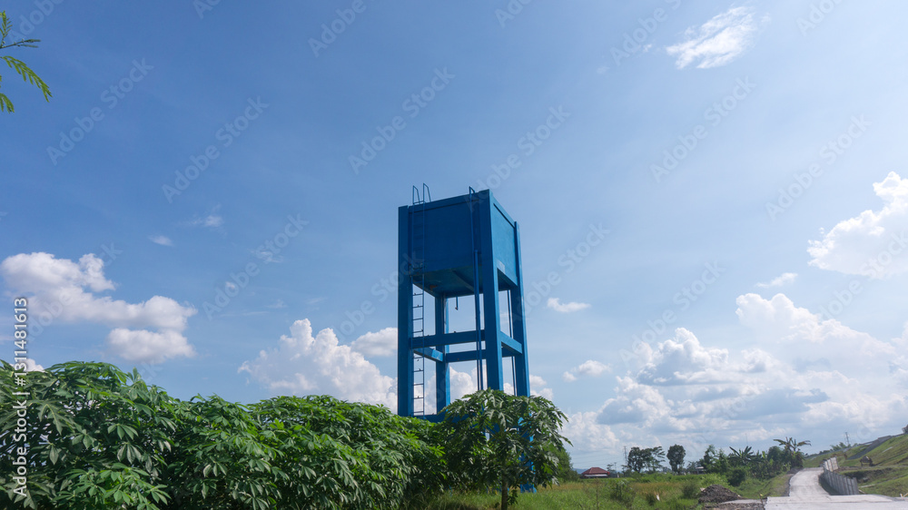 A blue elevated water tank stands against a bright sky, surrounded by lush greenery and a rural landscape. A vital water supply structure in a countryside setting with a clear blue sky. A blue elevated water tank stands against a bright sky, surrounded by lush greenery and a rural landscape. A vital water supply structure in a countryside setting with a clear blue sky.