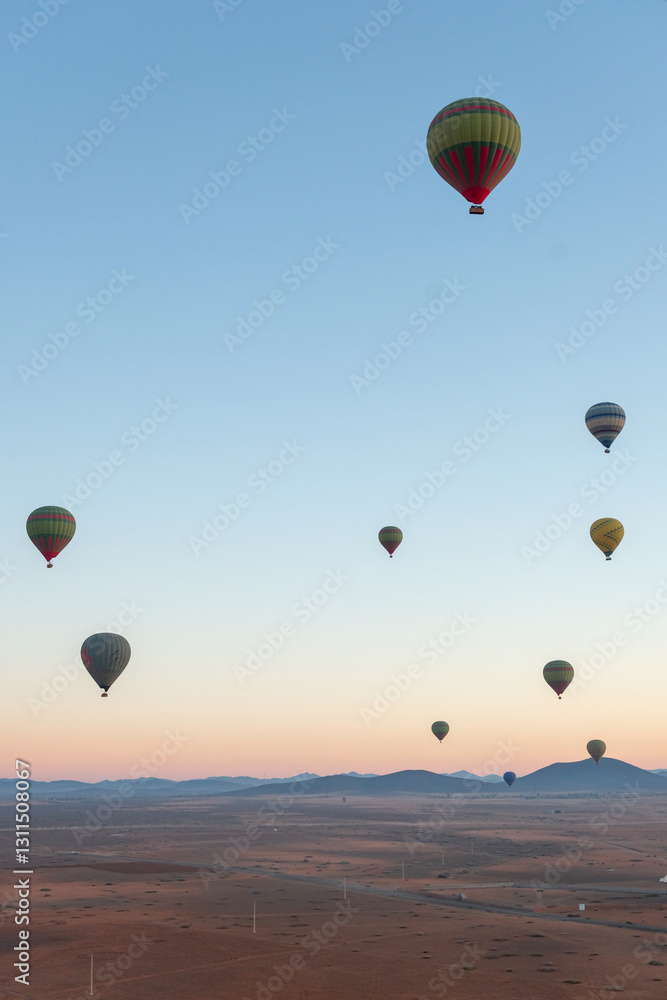 Fototapeta premium Hot air balloons float above an open landscape at sunrise, with mountains in the background. The sky transitions from deep blue to warm hues of dawn.