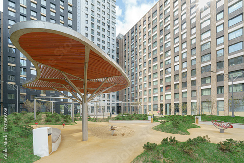 A park with a wooden pavilion and benches. The pavilion is covered in wood and has a curved roof. The park is surrounded by buildings, with one of them being a large apartment building.
