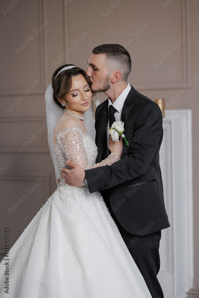 A bride and groom are hugging each other in a wedding photo. The bride is wearing a white dress and the groom is wearing a black suit. Scene is happy and loving
