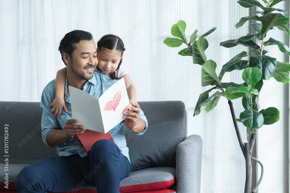 Little daughter kid making surprise present for her father for father's day or birthday, giving dad a handmade card with heart and reading together on sofa at home. Asian family celebrating holiday Little daughter kid making surprise present for her father for father's day or birthday, giving dad a handmade card with heart and reading together on sofa at home. Asian family celebrating holiday