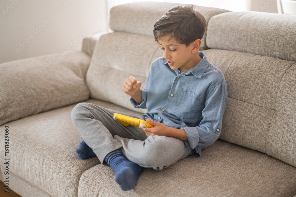 Concentrated child sitting on comfortable sofa with legs crossed, holding yellow handheld video game console and playing exciting game in cozy living room Concentrated child sitting on comfortable sofa with legs crossed, holding yellow handheld video game console and playing exciting game in cozy living room