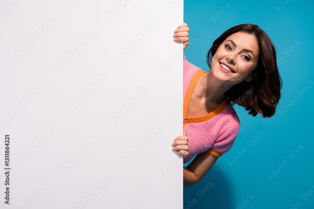 Cheerful young woman smiling playfully behind a blank display in colorful casual outfit Cheerful young woman smiling playfully behind a blank display in colorful casual outfit