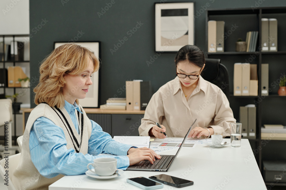 Obraz premium Portrait of two young businesswomen focused on work one girl typing on laptop keyboard with Asian colleague reviewing financial reports in office