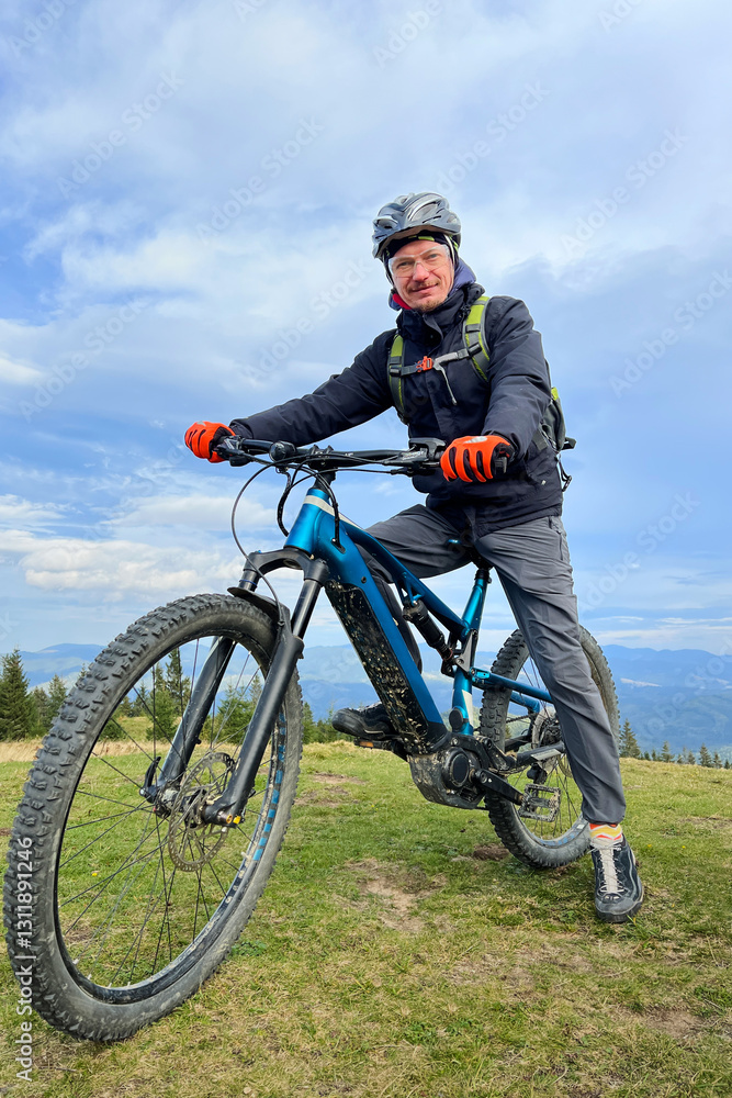 Cyclist man riding electric bike outdoors. Portrait of male tourist resting on grassy hill, enjoying beautiful mountain landscape, wearing helmet and backpack. Concept of active leisure. Cyclist man riding electric bike outdoors. Portrait of male tourist resting on grassy hill, enjoying beautiful mountain landscape, wearing helmet and backpack. Concept of active leisure.