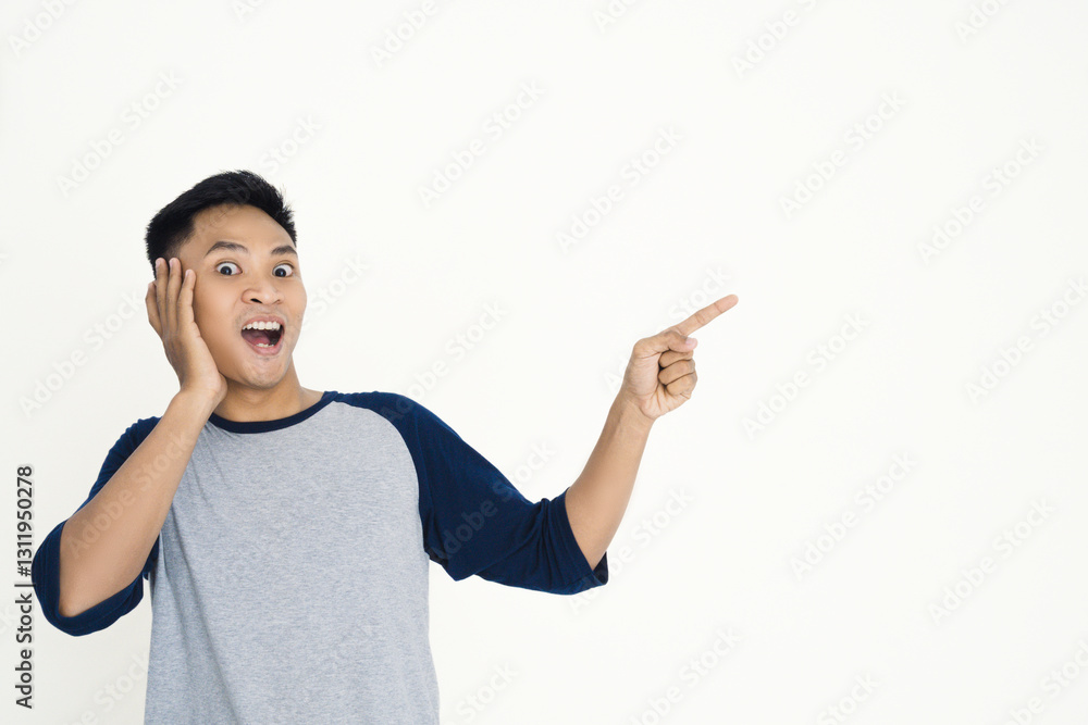 Young Asian man showing excitement while pointing to the side, wearing a casual gray and blue shirt and expressing emotions of amazement and joy against a plain white background. Young Asian man showing excitement while pointing to the side, wearing a casual gray and blue shirt and expressing emotions of amazement and joy against a plain white background.