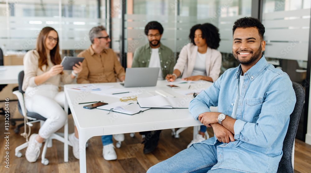 Fototapeta premium Young professional in denim shirt smiling confidently while sitting at a business meeting with a diverse team in a modern office space.