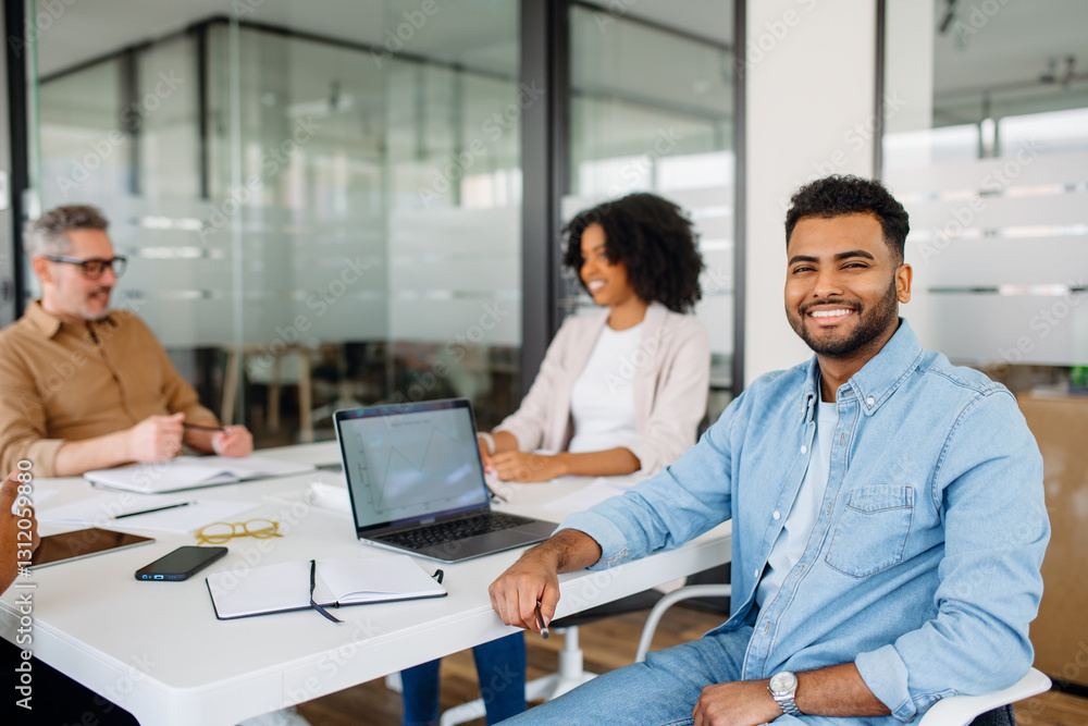A confident young professional in a denim shirt smiling at the camera while sitting in a bright and modern office space. His positive energy reflects teamwork and a friendly work environment. A confident young professional in a denim shirt smiling at the camera while sitting in a bright and modern office space. His positive energy reflects teamwork and a friendly work environment.