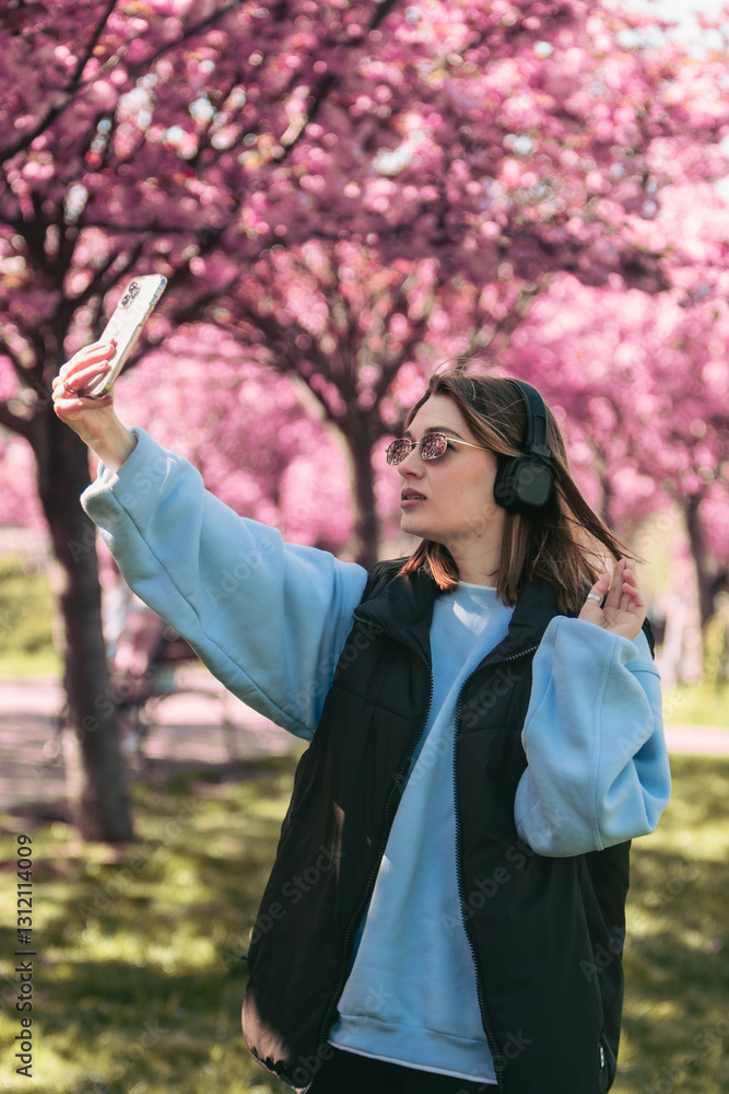 happy woman listening music in headset outdoors taking selfie with sakura on background