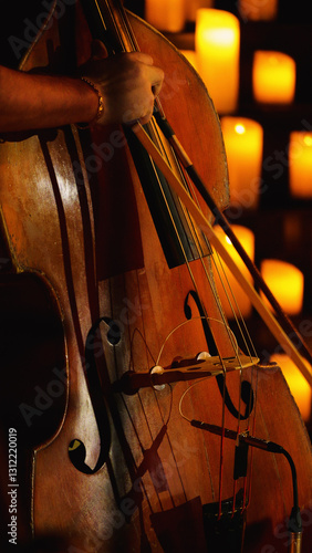 A close-up of a double bass being played in front of a wall of lit candles. The background is blurred and orange.