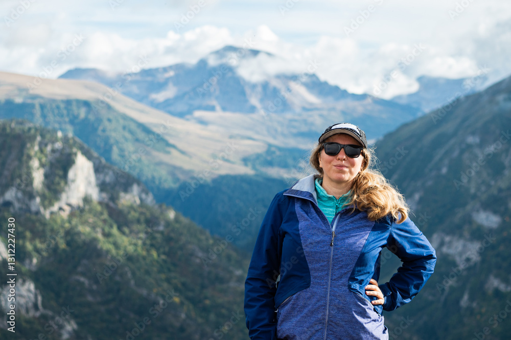 Girl tourist in sunglasses, baseball cap and windbreaker standing against the background of mountains. The slopes of the mountains are covered, green, and the main peak in clouds. Mountain hiking. Girl tourist in sunglasses, baseball cap and windbreaker standing against the background of mountains. The slopes of the mountains are covered, green, and the main peak in clouds. Mountain hiking.