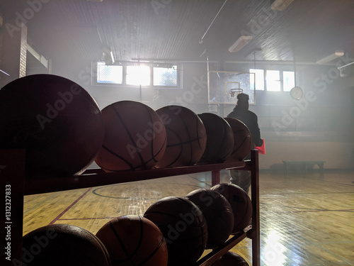 A quiet gymnasium setting lit by soft natural light, featuring basketball equipment and a shadowy figure in the distance, evoking themes of sports, solitude, and inspiration.