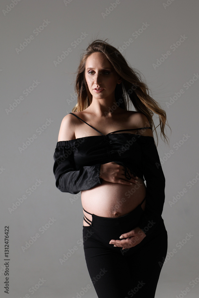 Portrait of attractive pregnant woman in black costume, trousers, top, studio photoshoot. Fashion photo of pregnancy. Young pretty girl with long hair touching her belly. Isolated, grey background Portrait of attractive pregnant woman in black costume, trousers, top, studio photoshoot. Fashion photo of pregnancy. Young pretty girl with long hair touching her belly. Isolated, grey background