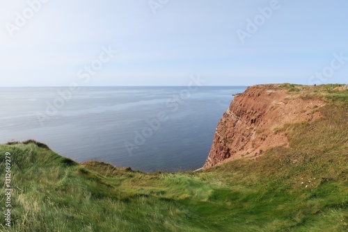 Vom Klippenrandweg auf der deutschen Hochseeinsel Helgoland sind die Felsen aus Buntsandstein, Gras darauf und die Nordsee unten zu sehen. Die See ist sehr ruhig an diesem Sommertag.