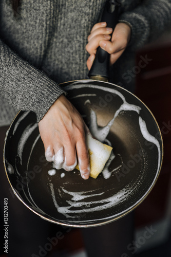 Diligent woman housewife washes, cleans with yellow sponge and detergent with foam dirty teflon round frying pan, dishes in kitchen. Close-up photo, concept of care and cleaning.