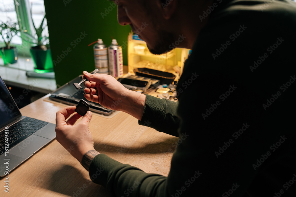 Focused technician repairing smartwatch battery using tweezers for precise component extraction and replacement in dark workshop sitting at desk with laptop computer. Focused technician repairing smartwatch battery using tweezers for precise component extraction and replacement in dark workshop sitting at desk with laptop computer.