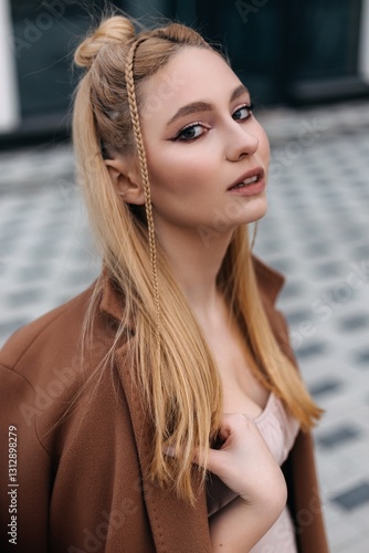Professional fashion model showcasing a trendy hairstyle with braids, wearing a stylish brown coat, posing confidently outdoors with a blurred urban background
