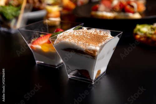 A portion of tiramisu cake in a plastic cup, sprinkled with chocolate chips. The dish consists of delicate white cream and biscuits. A small sweet dessert for an appetizer on a black background.