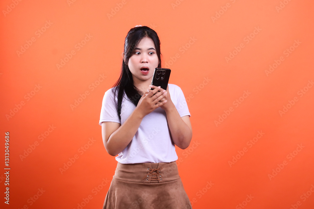 A young woman asia wearing a white shirt and brown skirt is looking at her phone with a surprised expression. Her mouth is open and her eyes are wide isolated on an orange background. A young woman asia wearing a white shirt and brown skirt is looking at her phone with a surprised expression. Her mouth is open and her eyes are wide isolated on an orange background.