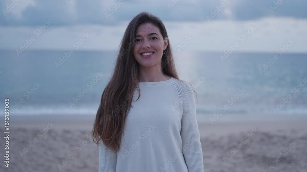 A young woman with long brown hair smiles gently while standing on the beach, with the ocean and a soft, cloudy sky in the background. Dressed in a cozy white sweater, she touches her hair A young woman with long brown hair smiles gently while standing on the beach, with the ocean and a soft, cloudy sky in the background. Dressed in a cozy white sweater, she touches her hair