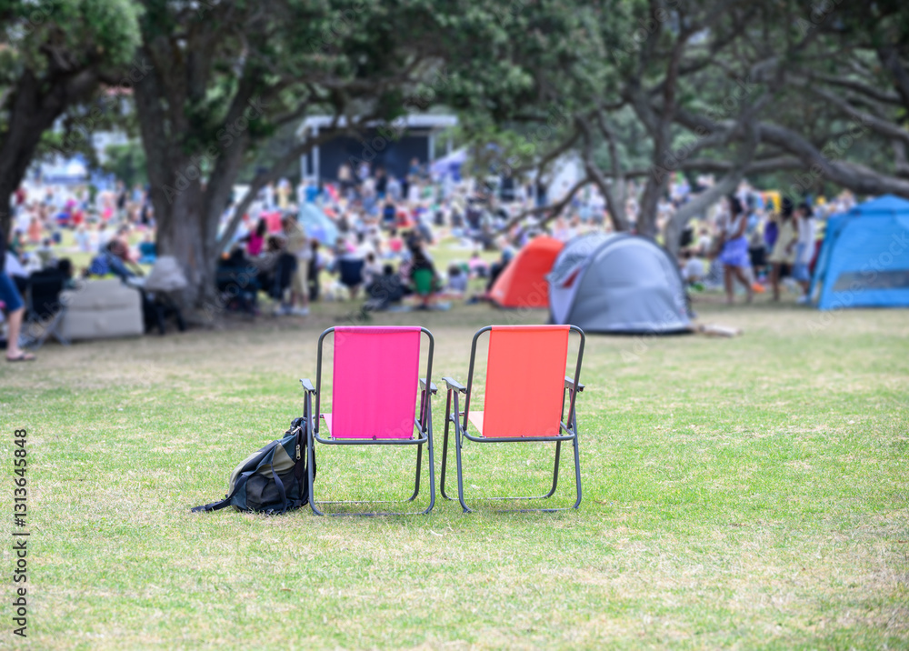 Two empty camping chairs, unrecognizable crowds listen to the music concert in the park. Auckland.