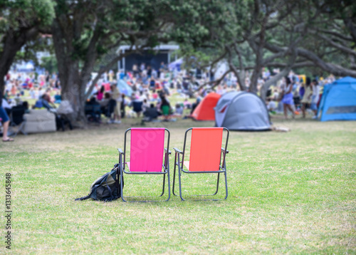 Obraz na płótnie Two empty camping chairs, unrecognizable crowds listen to the music concert in the park. Auckland. w salonie