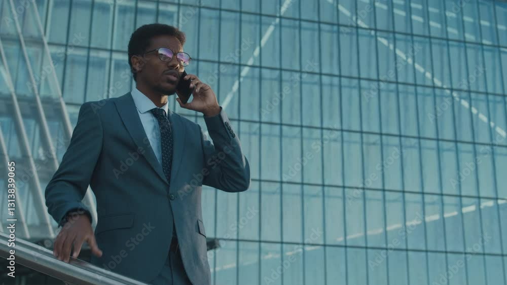 Well-groomed African American businessman in suit and glasses, having intense mobile phone call, standing on elevated platform outside modern, grid-patterned glass building Well-groomed African American businessman in suit and glasses, having intense mobile phone call, standing on elevated platform outside modern, grid-patterned glass building