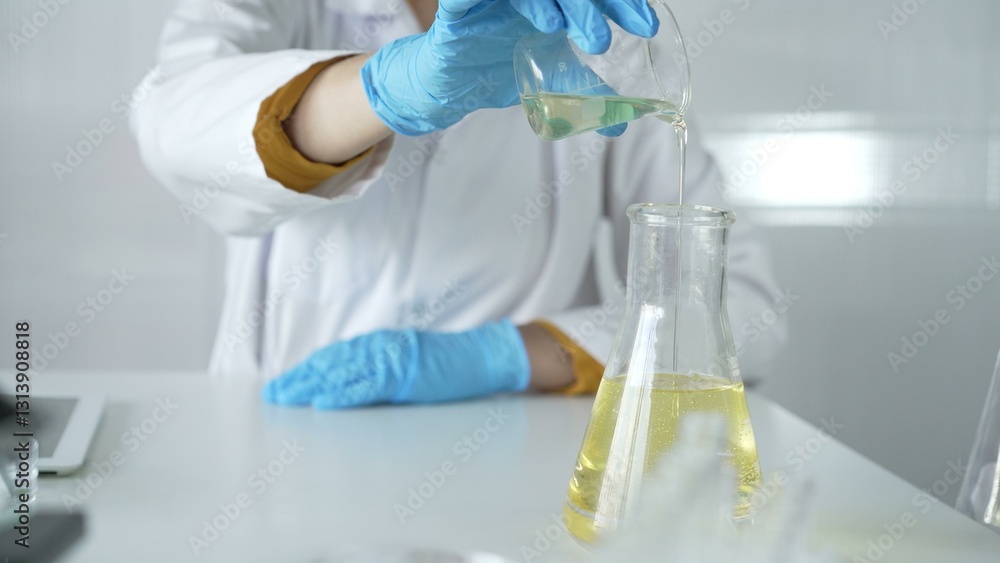 Fototapeta premium A scientist, wearing a lab coat and blue protective gloves, is pouring a yellow liquid from one beaker to another in laboratory, close up. Medicine and science concepts