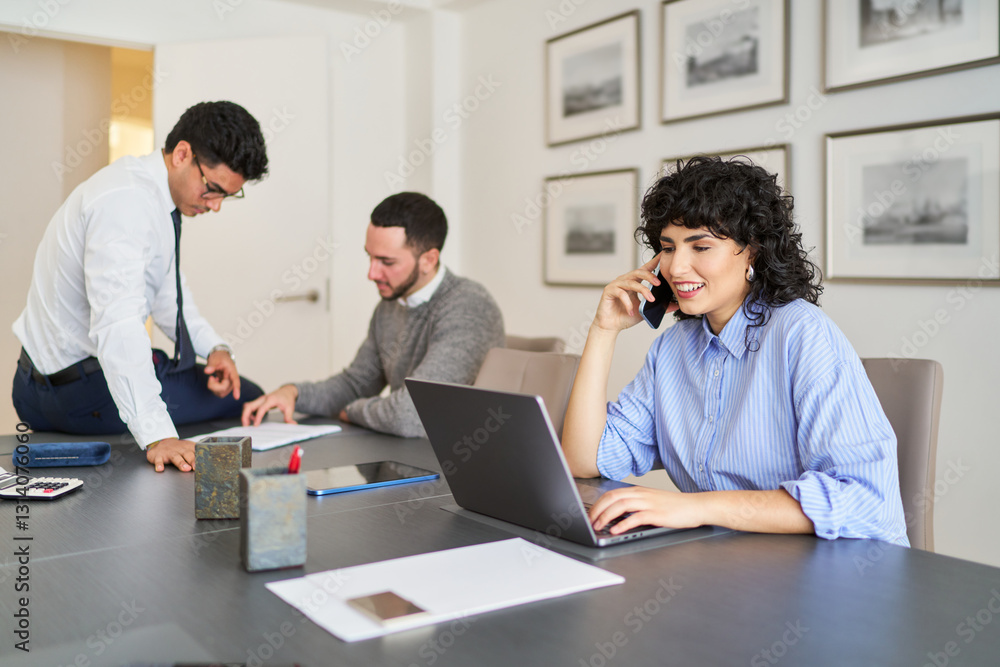 Businesswoman talking on phone and working on laptop in office with colleagues Businesswoman talking on phone and working on laptop in office with colleagues