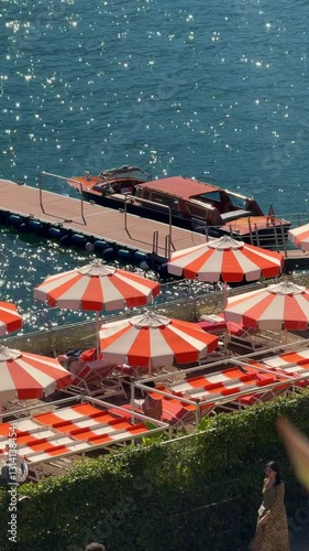 classic wooden private boat swinging on the waves near wooden pier and private beach club with traditional orange white sun umbrella of famous Como lake in north Italy