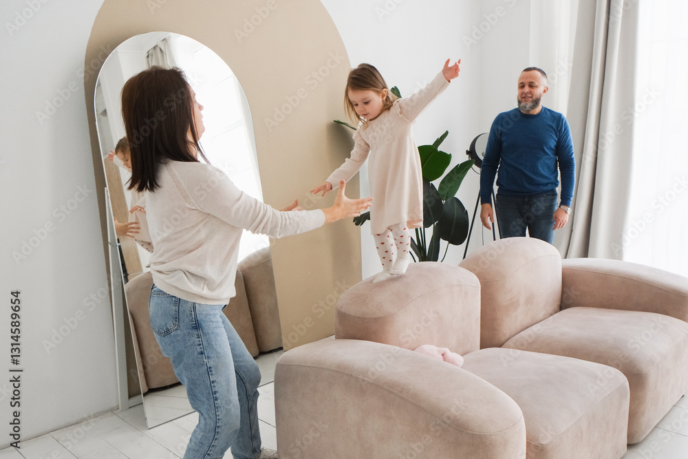 Obraz premium Mother encourages her daughter to balance on soft furniture in a bright living room. Father observes with pride, showcasing the warmth and love of family interactions and fun time