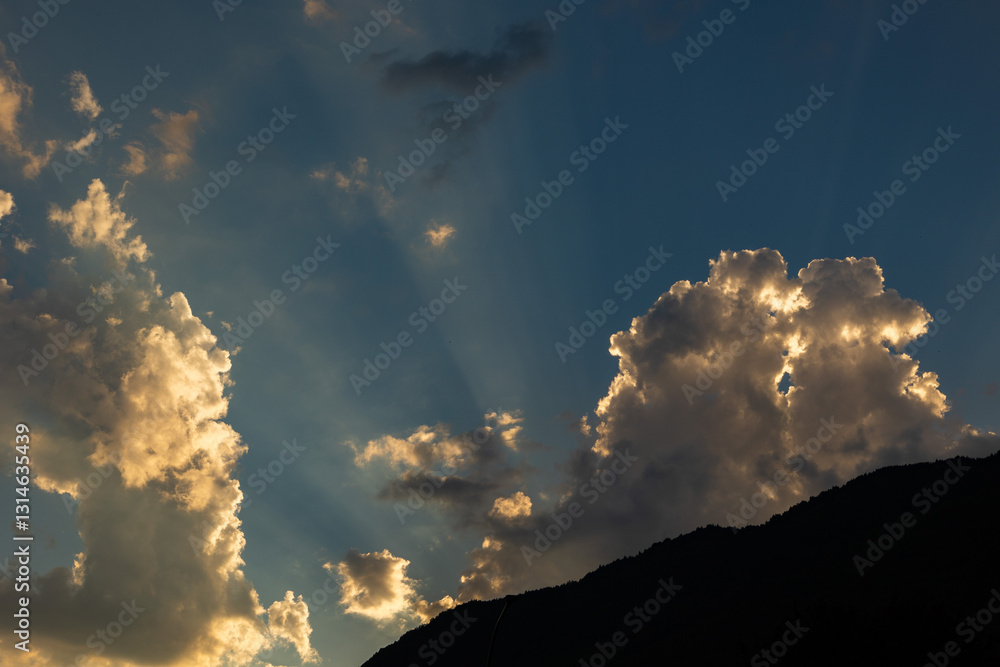 luce calda del sole al tramonto che illumina le nuvole ed il cielo di arancione da dietro un'alta catena montuosa, in estate