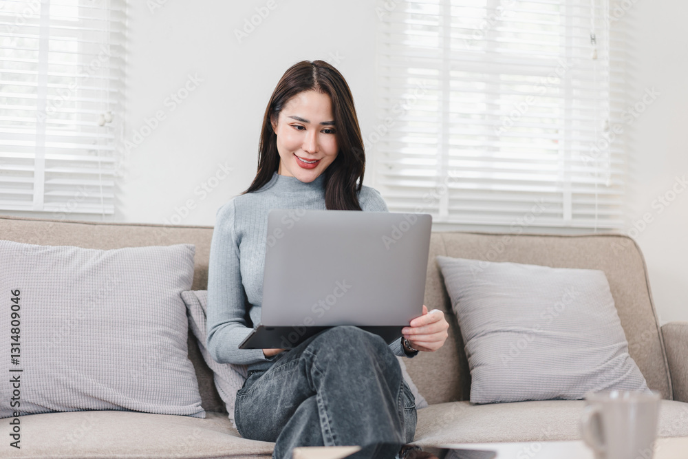 Asian Businesswoman Sitting on Sofa, Smiling While Using Laptop in Bright Living Room. Asian Businesswoman Sitting on Sofa, Smiling While Using Laptop in Bright Living Room.