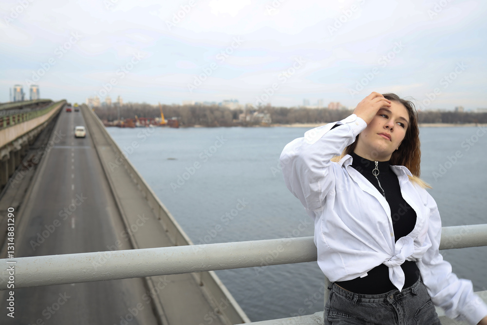 Young woman poses gracefully on a bridge overlooking a serene river with city skyline in the background during a cloudy day Young woman poses gracefully on a bridge overlooking a serene river with city skyline in the background during a cloudy day