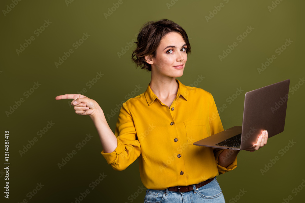 Φωτογραφία Young professional woman pointing confidently while holding a laptop, wearing stylish yellow blouse against khaki green background