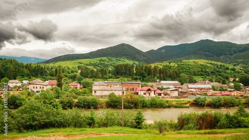 Obraz na płótnie Picturesque village nestled in green hills by a river under a cloudy sky offers a tranquil landscape. w salonie