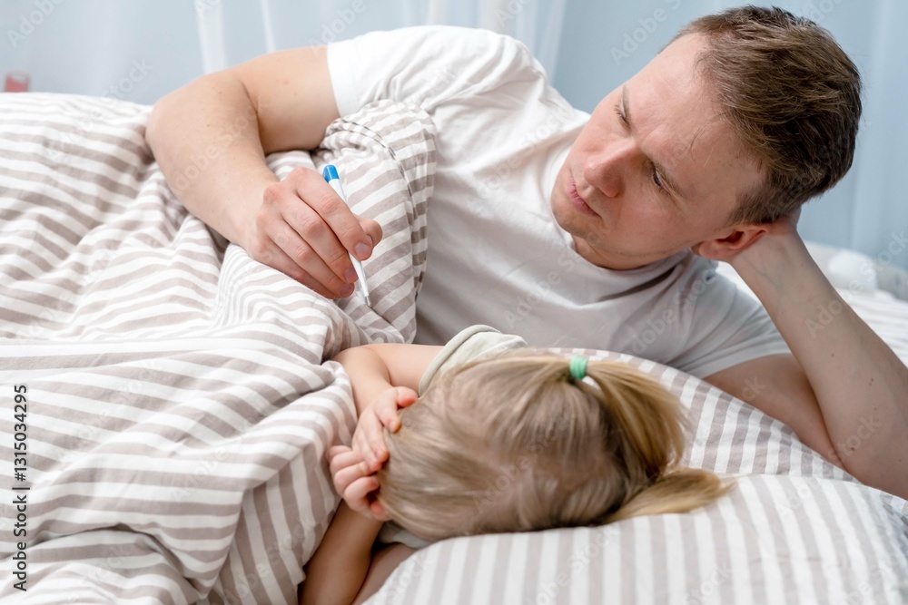 A father lies in bed with a sick child, a girl, and measures her body temperature with a thermometer in her armpit. A father lies in bed with a sick child, a girl, and measures her body temperature with a thermometer in her armpit.
