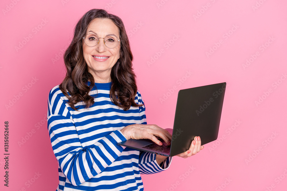 Naklejka premium Smiling mature woman in striped sweater holding laptop in front of pink background for advertising or lifestyle promotion