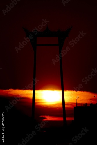 Silhouette of the Giant Swing in Bangkok, Thailand, against a stunning sunset sky. The scene is bathed in deep oranges and reds, creating a contrast.   