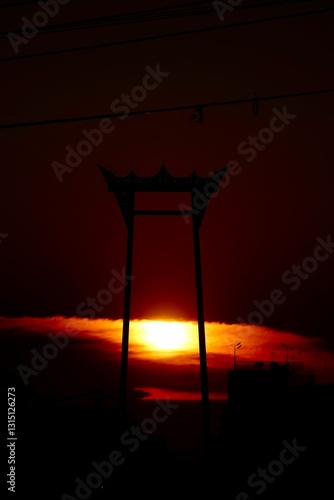 Silhouette of the Giant Swing in Bangkok, Thailand, against a stunning sunset sky. The scene is bathed in deep oranges and reds, creating a contrast.   