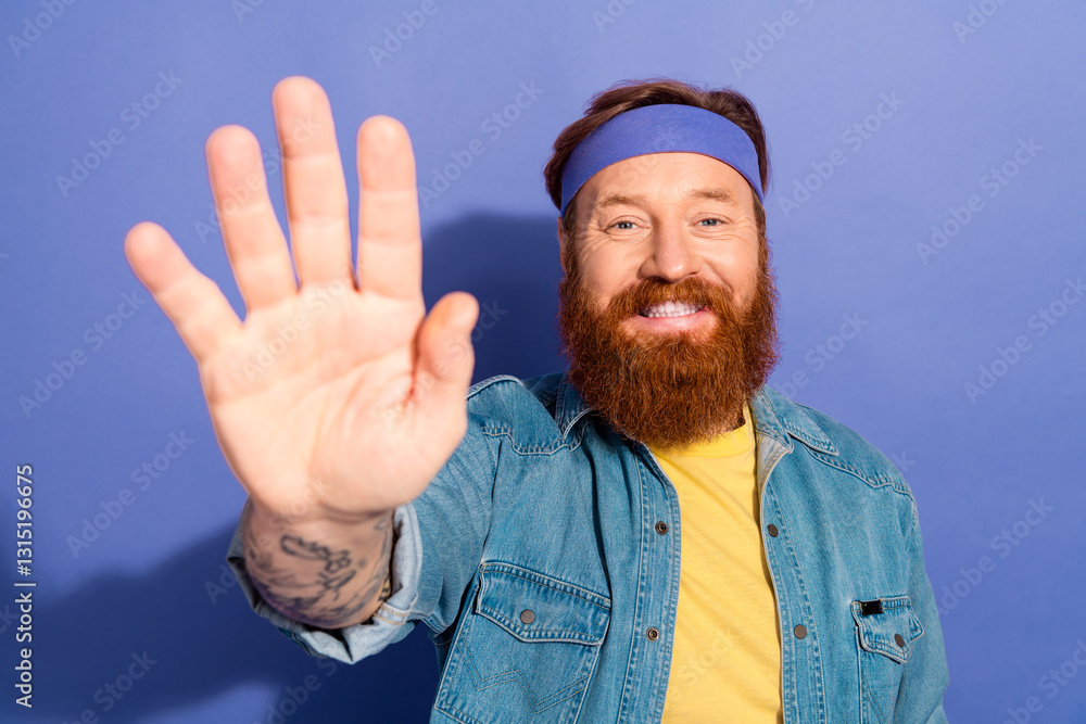 Naklejka premium Portrait of a cheerful bearded man wearing a denim jacket and headband against a vibrant purple background, giving a happy wave