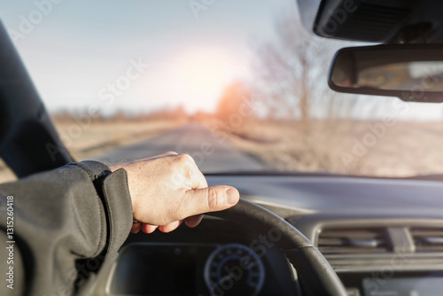 A person's hand on a car's steering wheel, driving along a forested road. The interior of the vehicle with a dashboard and rearview mirror. Forested road and sunset.