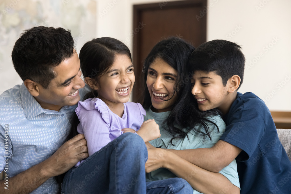 Beautiful carefree Indian parents play with two adorable kids, family having fun sitting on sofa tickling each other and laughing, exuding happiness, enjoy playtime together on weekend leisure at home Beautiful carefree Indian parents play with two adorable kids, family having fun sitting on sofa tickling each other and laughing, exuding happiness, enjoy playtime together on weekend leisure at home