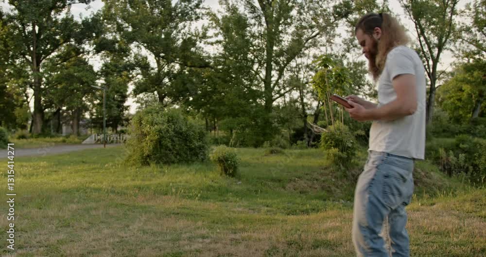 A bearded man in jeans and white t-shirt walks through park using tablet. Modern professional in nature, finds peace and harmony using technology. A bearded man in jeans and white t-shirt walks through park using tablet. Modern professional in nature, finds peace and harmony using technology.