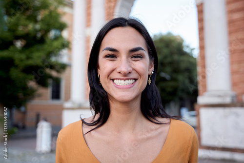 Portrait of a cheerful young latin woman with long dark hair and golden earrings, wearing an orange shirt and smiling broadly while standing in front of an urban archway Free Download Portrait of a cheerful young latin woman with long dark hair and golden earrings, wearing an orange shirt and smiling broadly while standing in front of an urban archway - GFXLoad