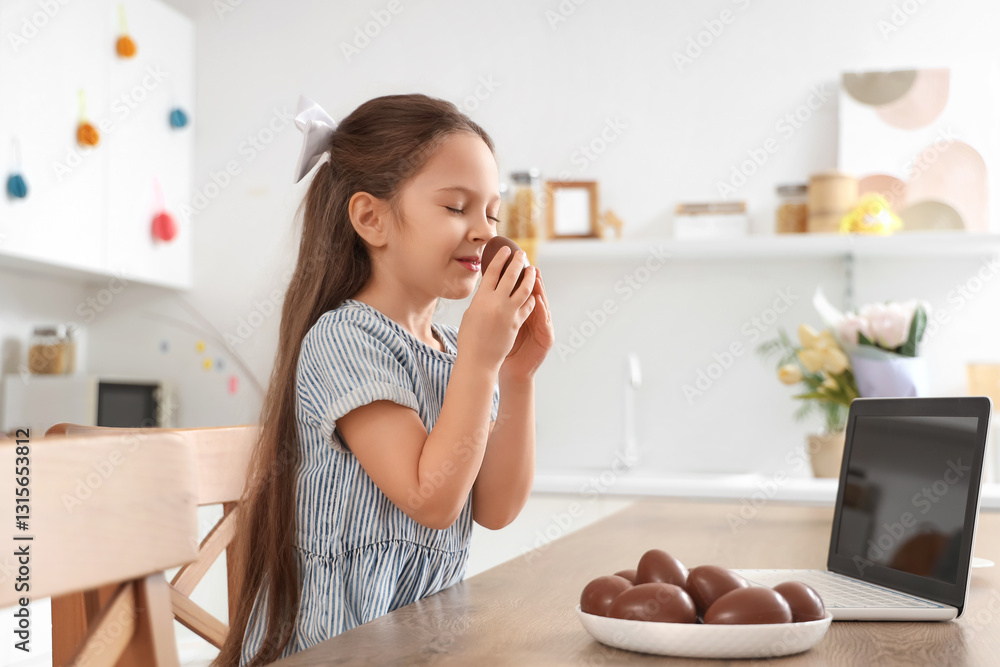 Cute little girl with plate of sweet chocolate eggs and modern laptop at table in kitchen. Easter celebration