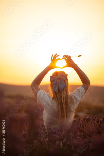 Back view of  beautiful girl  with bow in long hair, a woman strolls trough a lavender field at sunset. The golden light bathes the landscape, casting a warm blow on the flowers