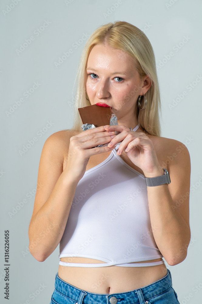 Naklejka premium Attractive young blonde eating a chocolate bar. She is wearing a white top and blue jeans on a white background. Copy space