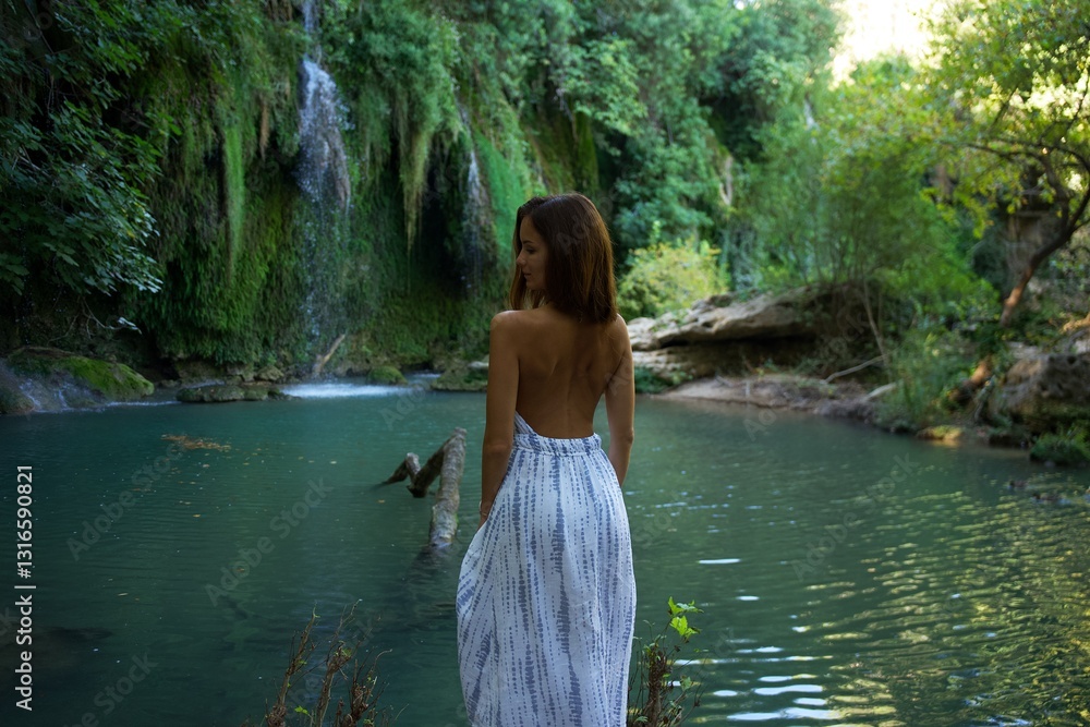 Fototapeta premium A woman in a flowing backless dress stands by a turquoise river, gazing at a cascading waterfall, surrounded by lush greenery and peaceful natural beauty. 